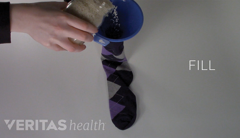 Rice being poured into a sock to create a homemade heating pack. Play button overlayed to access How to Make a Moist Heat Pack Video